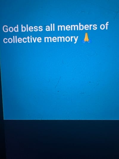 The scene, located in the city of Nnobi, Nigeria, presents a close-up view of a digital display screen. The upper two-thirds of the screen display a uniform medium-blue background. White, sans-serif text is centered on this blue field, reading: "God bless all members of collective memory". Immediately following the word "memory" is a yellow-orange praying hands emoji (🙏). The screen's surface shows multiple faint, linear abrasions and specks of dust, consistent with a physical display monitor. The bottom third of the image is occupied by a dark, unidentifiable plane, possibly a surface below the screen or the screen's lower bezel in shadow. No individuals, specific actions, or further objects are visible.