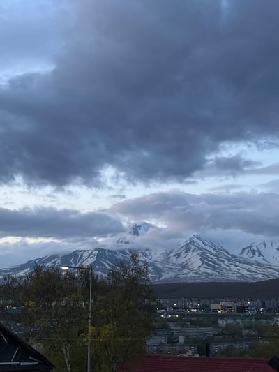 The image shows a panoramic view of Petropavlovsk-Kamchatsky, Russia, with the distinctive snow-capped volcanoes of the Kamchatka Peninsula in the background. The sky is filled with dramatic, dark clouds, suggesting either an approaching storm or the aftermath of one. In the midground, a town or city is visible, with buildings and trees. A street lamp stands prominently in the foreground, illuminating a section of the trees. The overall mood is somber and majestic, dominated by the imposing natural landscape.