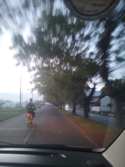 This is a motion-blurred photograph taken from inside a vehicle, likely a car, looking out at a road scene. The primary subject is a person riding a motorcycle down the road. The motorcycle's taillight is illuminated, suggesting it's either dusk, dawn, or nighttime. Trees line the right side of the road, their forms elongated and indistinct due to the motion blur. In the background, there are buildings and some vegetation. The sky appears overcast or hazy. The time of day seems to be either early morning or late evening, given the low light and the illuminated motorcycle taillight. The location context is Banda Aceh, Indonesia. There is no visible text in the image. The blur suggests the vehicle was moving when the photo was taken, capturing a sense of travel or transit.