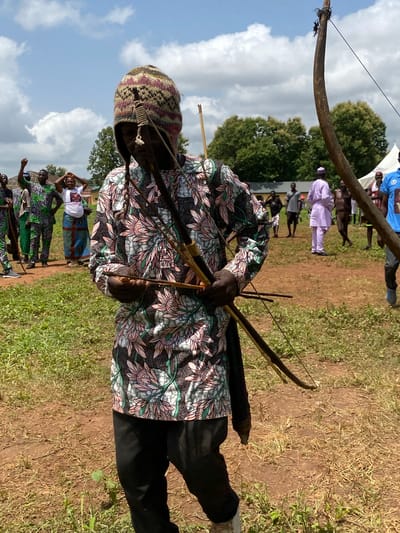 An African man in Maigizo, Nigeria, stands on a patch of dry dirt and grass holding a bow and arrows. He wears a patterned floral shirt, black pants, and a knit hat. Behind him are other people and a wooden structure. The sky is visible with some clouds.