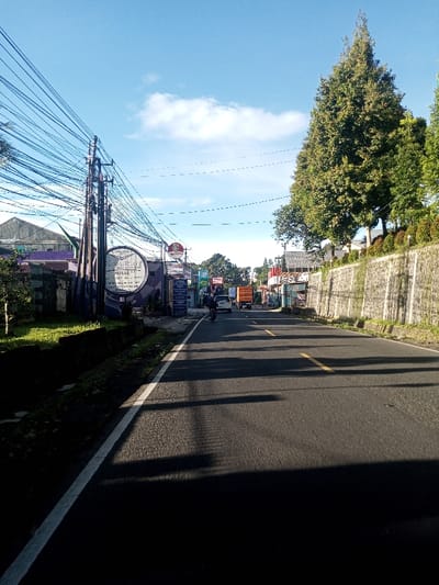 This image depicts a street in Khouathineung, Laos, under a clear blue sky with sparse white clouds. The asphalt road occupies the central and lower portions of the frame, featuring a solid white line on its left edge and dashed yellow lines indicating the center further ahead. Long, distinct shadows extend from the left side across the road surface.
On the left side of the road, multiple utility poles support a dense array of overhead wires. Below these, a narrow strip of grass is bordered by a low wall. Commercial structures line this side of the street; one building features a prominent circular sign displaying "elora cafe" and "HOTEL MARGONO." Another visible sign is a red circle with "TOKO."
The right side of the road is defined by a stone retaining wall, partially covered with climbing vegetation. Behind the wall, several tall, conical evergreen trees are visible, followed by additional structures in the distance.
Traffic moving away from the camera in the middle distance includes one individual operating a dark-colored motorcycle, followed by a white passenger vehicle and an orange truck.