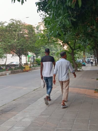 The image shows two men walking side by side on a sidewalk in Dhaka, Bangladesh, at the coordinates 23.801° N, 90.411° E. The man in the front is wearing a light blue shirt and beige pants, while the man behind him is wearing a white shirt with a black stripe on the chest and blue jeans. They are both walking in the same direction, away from the camera. There is a road on the left side of the sidewalk and a row of trees on the right. The sidewalk is made of concrete and has a yellow stripe running along its edge. There are also some yellow bricks embedded in the sidewalk. There is a light pole in the background and a white car parked on the road. The weather is clear and the sky is blue.