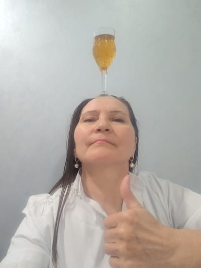 A medium shot shows a woman posing with a glass of beer or cider balanced on her head. She gives a thumbs-up with her right hand. The woman has medium-length, dark hair that appears wet. She wears a white button-down shirt and small, round earrings. The background is a plain, light-colored wall. There is no information suggesting the location is Sarapul, Russia.