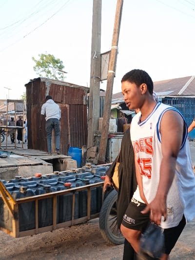 The image depicts an outdoor street scene in Gwagwarwa, Nigeria. In the immediate foreground on the right, a person with short dark hair, wearing a white sleeveless basketball jersey with red "INGS 55" text and an NBA logo, and black shorts, walks from right to left. A black garment is draped over their left arm, and a dark object is held in their left hand.
Immediately to the left of this person, a wooden pushcart is positioned, laden with approximately eighteen dark-colored rectangular containers, each featuring an orange cap. In the mid-ground, a male subject wearing a light grey hoodie and dark trousers is observed from behind, stepping onto a raised wooden platform that leads to a structure made of weathered, rusted corrugated metal sheets. A blue plastic barrel is situated near the base of this structure.
Two utility poles, one concrete and one wooden, stand vertically in the center-right, supporting overhead wires. The background features various buildings with corrugated metal roofs, additional pedestrians, and a parked motorcycle on the far left. The ground throughout the scene is unpaved dirt. The sky is clear and bright.