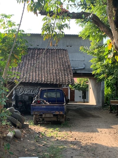 The image depicts a scene in what appears to be a residential or workshop area in Tanjungsepreh, Indonesia. A blue pickup truck is parked on a dirt road. Behind the truck, a building with a tiled roof and a large banner with white lettering that reads "Dozag" is visible. To the right of the truck, there is an open passageway leading into another area with trees and a building. The environment is characterized by vegetation, a dirt ground, and a building that suggests a humid, tropical climate. The lighting indicates it is daytime, with shadows cast by the sun. There are no people visible in the image.
