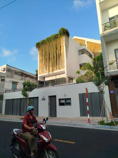 The image depicts a street scene in Ho Chi Minh City, Vietnam. A man wearing a red shirt and a light blue helmet is riding a red scooter on a dark asphalt road. Behind him are buildings, including a white, modern-looking house with vegetation cascading from its upper portion and a geometric screen facade. A street with grey roll-up garage doors, a striped pole, trees, and a neighboring white building with a balcony are also visible. The sky is blue with scattered clouds.