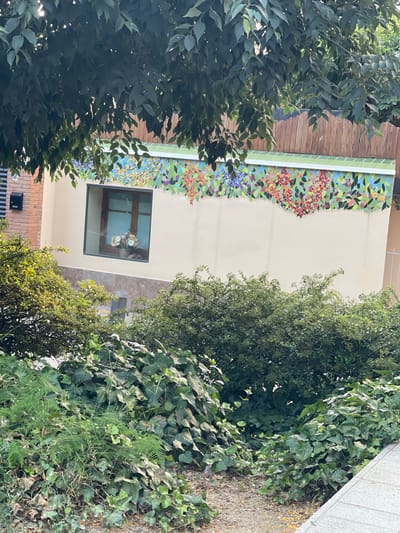 The image captures a section of a building's exterior in Santa Coloma de Gramenet, Spain, characterized by lush greenery and a vibrant mosaic. The building features a neutral-colored facade adorned with a colorful mosaic depicting flowers and leaves along its upper section, below a band of green tiles. A rectangular window with wooden framing is visible on the facade, with a basket of flowers appearing to be placed inside. In the foreground, dense vegetation including ivy and other leafy plants creates a natural barrier, with a gravel path and a light-colored paved area to the right. The presence of a brick section of the building and a mailbox suggests an urban or suburban setting.