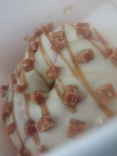 The image depicts a doughnut with white icing, caramel drizzle, and caramel cube toppings. The doughnut is situated inside a white box. The location is unknown.