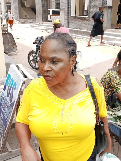 This image captures a daylight street scene in Ura Nta Umuarandu, Nigeria. In the foreground, a middle-aged Black female with dark braided hair and gold hoop earrings is prominent, facing slightly to the right of the frame. She wears a bright yellow v-neck top adorned with silver-colored embellishments and featuring cut-out shoulders. A black shoulder bag with a brown braided strap hangs from her right shoulder. To her left, a white and blue "POS" sign, advertising financial services, is visible on a wooden stand. To her right, another individual is partially visible, dressed in a patterned garment, attending to items, including green leaves, on a low surface.
In the midground, a black motorcycle with a green front wheel rim is parked behind the primary subject, facing left. Further to the left, a dark wheelbarrow is present. The background consists of a dusty, light-colored ground surface and various buildings constructed from bricks and concrete, some appearing to be under construction with exposed stairwells. Several other individuals are in motion; a male in a white shirt and red shorts walks away to the left, a male in a pink shirt and yellow baseball cap is partially obscured, and a male in a black top and a black skirt-like garment walks left, wearing sandals. A white utility van with blue and orange stripes is partially visible in the distant background. The scene suggests a busy, urban commercial area or market.