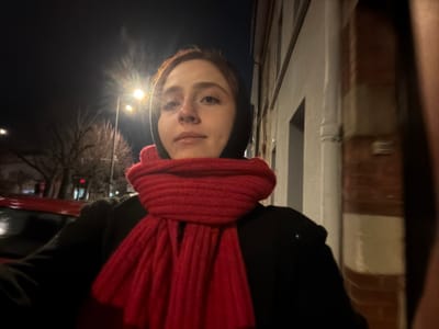 A person in a black coat and red scarf is shown at night. Streetlights illuminate the background, where trees and buildings are visible. The brick building is on the left. The person is centered in the frame, with their face clearly visible, looking toward the camera. The image is rotated.