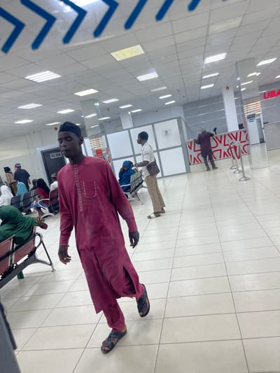 This image depicts the interior of a UBA bank branch in Zaria, Nigeria. In the foreground, a dark-skinned male wearing a maroon traditional tunic with embroidered patterns, matching trousers, and dark sandals is walking from left to right, his left foot mid-stride. His gaze is directed downwards and to his right.
In the midground, to the left, a row of metallic benches with red and green seating is partially visible, occupied by several individuals. A dark doorway marked "BULK ROOM" is present. To the right of the walking man, another dark-skinned male stands facing right, wearing a white long-sleeved collared shirt, light brown trousers, and light-colored sandals. He carries a dark-patterned crossbody bag on his right side. Behind him, a person in a blue head covering sits on a bench.
The background features bank counters with white and red patterned facades. A dark-skinned individual wearing a patterned shirt and dark trousers is standing at one of these counters, facing away from the camera, engaged in a transaction. Red stanchions with ropes are positioned in front of the counter. The "UBA" logo in red lettering is visible on a pillar or wall section to the far right. The environment is well-lit by numerous recessed rectangular fluorescent lights in a white tiled ceiling. The floor is also white-tiled with a grid pattern.