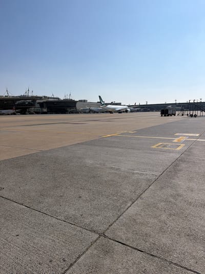 The image depicts an airport tarmac on a clear, bright day. The perspective is from the ground, looking towards the sky and down the length of the tarmac. The tarmac is concrete with yellow markings, including a box shape with a number inside, likely indicating a parking or gate position. Several airplanes are visible parked at the terminal in the background. The terminal building itself appears to be a modern structure with glass facades and multiple levels. Baggage carts and other airport equipment are also present on the tarmac. The sky is a pale blue, suggesting daytime. The overall scene is one of typical airport activity, with aircraft ready for departure or arrival. No people are prominently visible in the foreground or midground. The lighting and clear sky indicate it is likely midday.