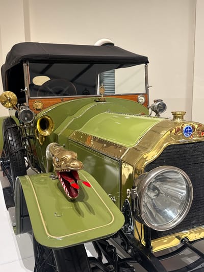 The image shows a vintage green and gold vehicle, possibly an antique car, likely on display in an indoor setting. It features distinctive brass elements, including a snake-shaped horn and ornate detailing around the radiator. The vehicle has a black soft-top and spare tire mounted on the front. The environment appears to be a museum or exhibition space, with neutral walls and lighting. The location is unknown but has no information from South Africa.