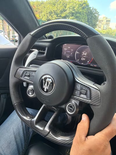 The image is an indoor shot taken from the perspective of a driver inside a car, specifically a Maserati based on the emblem on the steering wheel. The focus is on the car's dashboard and steering wheel, which features carbon fiber accents. The driver's hand is visible on the steering wheel, and their leg in blue jeans is also in the frame.

The digital dashboard displays various information, including trip data (642.0 km, 4.2 km/l average, 44 km/h), current time (08:59, 14:21), odometer reading (017839 km), current speed (34 km/h), transmission mode (PNDM - D2), and remaining distance (31 km). Controls for cruise control (RES+, CAN-, SET+) and audio/communication (phone button, microphone button, VOL A, VOL V) are visible on the steering wheel. A prominent "START STOP" button and a "LAUNCH" button are also present.

The background outside the car reveals a bright, sunny day with lush green trees and buildings, suggesting an urban environment. The presence of parked cars and a pedestrian crossing in the background hints at a street setting within a city.

Based on contextual information provided in the prompt, the scene is located in Givatayim, Israel.