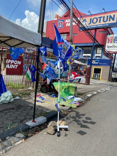 The scene shows a street in Cimahi, Indonesia, located at -6.891 latitude and 107.557 longitude. A white metal stand with wheels is positioned on the sidewalk. It is holding a collection of small blue and white flags, predominantly in a radial pattern, and a green and white cloth is at the base of the stand. The flags are primarily blue with white lettering. The flags are attached to the stand by strings, and some of the strings are connected to small, white plastic clips. The stand is facing the street, which has a grey asphalt surface and a concrete curb separating the street from the sidewalk. To the left of the stand, there is a grey sidewalk with a concrete curb. A white tent is located on the sidewalk, and there is a metal fence behind the tent. Behind the fence, there are buildings painted in red, blue, and white. The building on the far right is adorned with a yellow sign with the word "JOTUN," while the building next to it has the word "NILAM" written on it. The building's exterior indicates it is a paint store. The sign on the building on the right reads "SINAR TERANG ABADI Cimahi." There are power lines running over the buildings. The sky is partly cloudy, and the sun is shining.