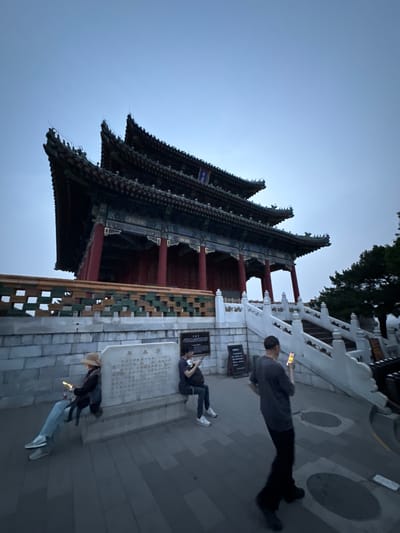 The image shows a Chinese building with multiple roofs, likely a traditional temple or palace structure, located in the Xicheng District of China. Several people are present, with some looking at their phones. The environment appears to be a paved area surrounding the building, potentially a public space.