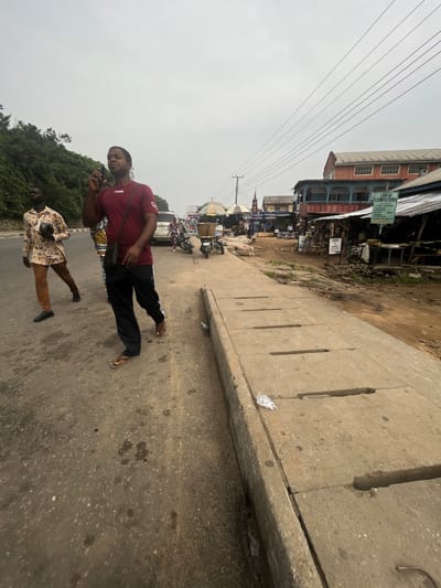 This image depicts a street scene in Ikot Ekpene, Nigeria, under an overcast sky. The foreground features a worn asphalt road with visible aggregate and scattered dark stains, running parallel to a concrete drainage channel on the right. This channel is covered by rectangular concrete slabs with slotted openings; a piece of white plastic litter is visible on one slab.

Two adult males are walking on the asphalt from left to right. The man in the immediate foreground wears a maroon short-sleeved t-shirt with "mess" printed on it, dark track pants with white side stripes, and brown flip-flops. He carries a small black shoulder bag and holds a black smartphone to his right ear, appearing to be engaged in a call or listening. Behind him, to the left, another man walks wearing a light-colored, patterned long-sleeved shirt, brown trousers, and dark shoes. He carries a dark, possibly spherical, object in his left hand.

To the left of the road, a densely vegetated hillside rises steeply. On the right side of the street, beyond the drainage, is a dirt shoulder leading to a series of commercial establishments. These include open-air market stalls with corrugated metal roofs and striped umbrellas. Signs are visible, notably "LENY-D COMPUTERS" offering typing, printing, lamination, and internet services, and "FOOD IBIAD PEPPER SHOP." Several motorcycles are parked or positioned near these stalls.

Further down the street, multi-story buildings are present. One building features a red-orange upper facade with multiple windows and a blue-painted enclosed balcony structure below. Another building, partially obscured in the distance, shows a dark steeple, suggesting a church. Overhead power lines span the upper portion of the scene. Several other individuals are visible further down the street, near the market activity and a parked white SUV.