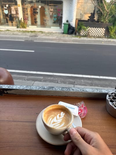A coffee cup with latte art rests on a saucer on a wooden table, with packets of sugar and a small container of sweetener nearby. A hand reaches for the cup. Adjacent to the table is a street view from Canggu, Indonesia, featuring a building with a store display window and a street with a white line marking a lane.
