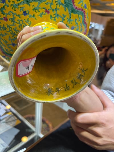 In this close-up, indoor shot, a person's hands are visible, holding an ornate yellow vase. The vase appears to be ceramic, with a slightly flared opening and a glossy yellow glaze. The exterior of the vase is decorated with a traditional Chinese motif featuring a green dragon. Inside the vase, there are faint black characters written vertically on the yellow glaze. A small, red-bordered white tag with some red text is attached to the inside of the vase's rim. The setting appears to be a retail environment, possibly an antique shop or gallery, suggested by the display shelves and lighting in the blurred background. The lighting is warm and artificial, indicating an indoor setting. The focus is on the vase and the hands holding it, highlighting the craftsmanship of the object and the interaction with it.