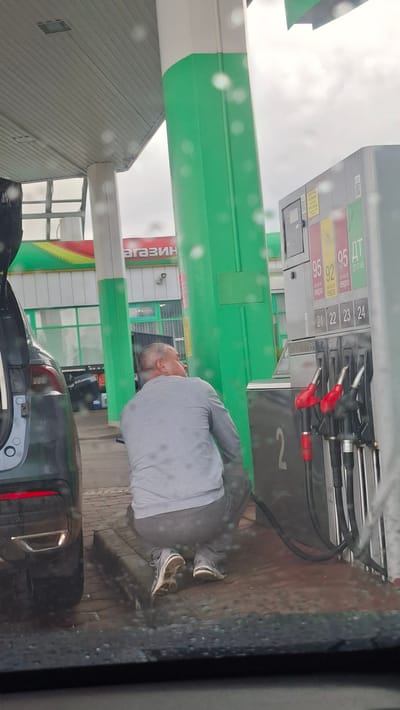 At a gas station in Smalyavichy, Belarus, a man wearing a gray sweatshirt and pants is squatting down next to a car. He appears to be in the process of fueling the vehicle, with a gas hose extending from the pump to the car. The gas pump has various fuel options displayed, including 95 and 92 octane gasoline, and diesel fuel. The scene takes place under a covered area of the gas station, with a green and white pillar visible. The ground is paved with reddish-brown bricks, and there are droplets of water on the surface and on the windshield from which the photo is taken, suggesting it is raining or has recently rained. The background shows a store with the word "МАГАЗИН" (MAGAZIN, meaning "shop" in Russian) visible on its facade.