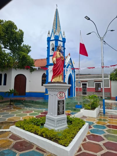 This is a full-shot, eye-level, outdoor, daytime photograph of a church entrance area in the city of Tacarigua, Venezuela. The scene is dominated by a statue of Jesus Christ in the foreground, standing on a pedestal. Behind the statue, the church building features a bright blue and white spire, a red-tiled roof, and arched doorways. A red and white flag flies from a pole to the right of the church. The ground in front of the church is paved with colorful, hexagonal tiles. There are no people visible in the image. The sky is overcast.