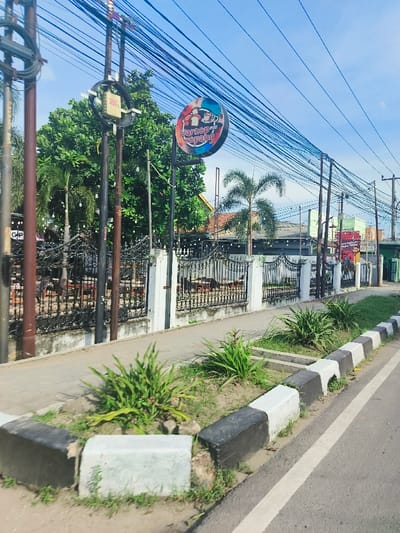 The image shows a sidewalk in Purwakarta, Indonesia, at coordinates -6.552, 107.437, with a black and white striped curb bordering a paved road. The sidewalk is lined with small, lush green plants and a black metal fence separates the sidewalk from a cafe on the left side of the image. The cafe is partially visible through the fence, showing a small wooden sign with the text "Warung Kopang" in red and white letters. Several power lines and electric poles run parallel to the road. The sky is clear and blue, and a large tree with green leaves fills the left side of the image. In the distance, another building with red and white letters is visible, although the text is not legible.