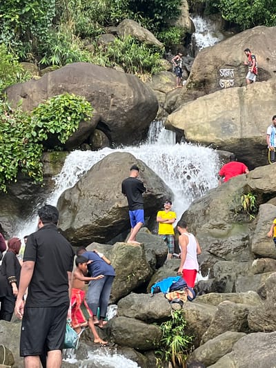 This image captures a lively scene at a waterfall in Jaflong, Bangladesh, a region renowned for its natural beauty and stone quarries. In the foreground, a man in a black t-shirt and shorts stands with his back to the camera, seemingly observing the activity. To his left, a woman in a black hijab and outfit is partially visible. Further into the scene, a group of young people are interacting around the waterfall and the rocky terrain. Two boys, one shirtless and wearing a red and green lungi, are being embraced by another person in a blue t-shirt and jeans.

The waterfall itself cascades down large boulders, creating a dynamic and refreshing atmosphere. Several individuals are positioned on the rocks, some wading in the water or sitting. One person in a bright yellow t-shirt and checkered shorts is visible near the base of a large rock. To the right, a person in a white tank top and red shorts stands on a rock, and further up on another rock, someone in a red t-shirt is seated.

Higher up, two individuals are on a large boulder with Bengali script inscribed on it. One is wearing a dark top and shorts, while the other has on a patterned shirt and shorts. Another person in a white and blue soccer jersey stands near them. The environment is lush with green vegetation clinging to the rocks and surrounding hills. The presence of multiple people suggests a popular tourist or recreational spot.