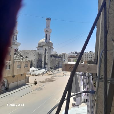 This image depicts the aftermath of destruction in the city of Khan Yunis, Palestinian Territories. The central focus is a damaged mosque, its dome partially intact, with significant structural collapse and debris scattered around. Adjacent buildings also show signs of damage, with rubble and broken walls visible.
In the foreground, on the left side of the image, is a partially obscured building with windows and air conditioning units. A sign with the letters "CA" and some symbols is visible on this building. Further down the street, two figures, an adult and a child, are walking away from the camera. The street itself is dusty and appears to be an urban environment with some tents or temporary structures visible in the background.
On the right side of the image, laundry is hanging on a clothesline, partially obscuring a concrete wall and some metal bars. Wires are also visible stretched across the scene against a clear blue sky. The overall impression is one of devastation and resilience, with daily life continuing amidst the ruins. The water mark "Galaxy A14" is present in the bottom left corner of the image.
