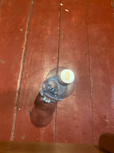 A top-down, medium shot captures a clear plastic bottle of water with a white and gold cap resting on a weathered, dark red wooden floor. The floorboards are wide and show signs of wear, with visible cracks and scuff marks. The bottle is positioned slightly off-center to the left, casting a distinct shadow to its right. The water inside the bottle is clear, and condensation is visible on the exterior, suggesting it is cold. The gold emblem on the cap appears to be a stylized depiction of a lion or other heraldic animal within a circular design. The overall scene is indoor, with a sense of an aged, rustic environment. There are no individuals present in the frame. The location is specified as Hoholeve, Ukraine.