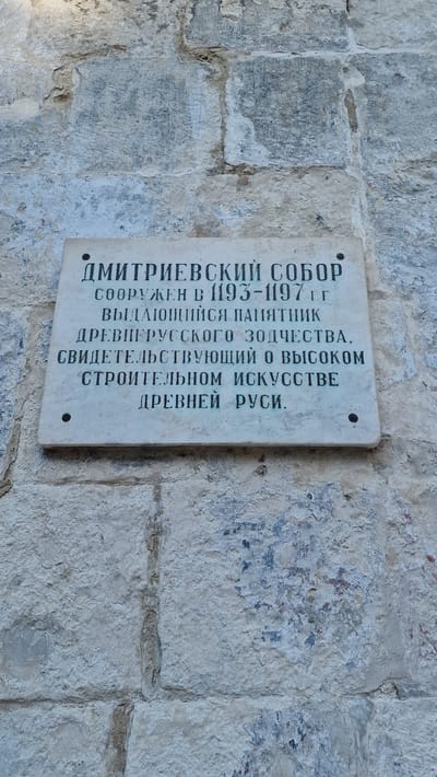 This is a vertical, eye-level, medium shot of a historical plaque mounted on a rough, light-colored stone wall. The plaque is off-white and rectangular with rounded corners, and it is oriented vertically, with the text running from bottom to top. The stone wall is textured, with visible cracks, uneven surfaces, and patches of what appears to be ancient plaster or paint in muted shades of blue and red. The lighting is bright, suggesting it is daytime.

The plaque bears an inscription in Russian. The text reads: "ДМИТРИЕВСКИЙ СОБОР СООРУЖЕН В 1193-1197 гг ВЫДАЮЩИЙСЯ ПАМЯТНИК ДРЕВНЕРУССКОГО ЗОДЧЕСТВА, СВИДЕТЕЛЬСТВУЮЩИЙ О ВЫСОКОМ СТРОИТЕЛЬНОМ ИСКУССТВЕ ДРЕВНЕЙ РУСИ." This translates to: "Dmitrievsky Cathedral Built in 1193-1197. An Outstanding Monument of Old Russian Architecture, Testifying to the High Building Art of Ancient Rus'." The plaque is attached to the wall with at least two dark, circular rivets or fasteners. The overall impression is one of historical significance and weathered resilience. The setting appears to be outdoors, likely an exterior wall of an ancient building.