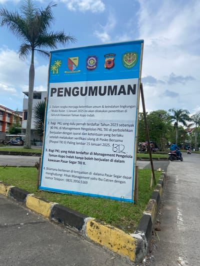 A white sign with the Indonesian word "Pengumuman" (Announcement) in large blue letters at the top is posted on a wooden pole. The sign is on a concrete sidewalk with yellow painted markings. There is a black and white curb separating the sidewalk from a grass lawn. There is a tree to the left of the sign. The sign is located at coordinates (-6.969, 107.552) which is in the Margaasih, Indonesia region. The sign contains the following announcement:
1. In the context of maintaining public order and environmental beauty, starting from January 1, 2025, the issuance of permits will be carried out in the entire Taman Kopo Indah area.
2. For PKLs who have registered in 2023, a total of 90 PKLs in the Management of PKL TKI will be allowed to operate after fulfilling the requirements and provisions that apply after carrying out verification at the Posko Bersama (Posko TKI II) at the latest on January 15, 2025.
3. For PKLs who are not registered in the Management of Taman Kopo Indah, are only allowed to trade within the Pasar Segar TKI II area.
4. If you wish to be accommodated in Pasar Segar, you can contact the Management Party, namely Ibu Catren with the telephone number 0831.3956.5169.
There is a handwritten "1312" on the sign. The sign also displays logos for the Polresta Bandung, the Satlantas Polresta Bandung, the Jawa Barat Regional Police, and the Silwandi Forestry and Land Affairs Office.