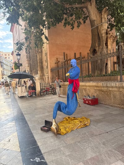 In Málaga, Spain, a street performer dressed as a genie is striking a pose on a tiled pedestrian walkway. The performer is wearing a blue jumpsuit with a red sash and a blue head covering, with their legs bent in an unusual position. A gold fabric drapes on the ground next to them, and a small open treasure chest rests near their feet. To the right, a red suitcase with wheels is visible. Behind the performer, a large, gnarled tree trunk looms, with a section of ornate metal fencing in front of it. A building with a warm, peach-colored facade and intricate architectural details is visible behind the tree.

In the background, several people are seated at an outdoor cafe, some with umbrellas providing shade. A bicycle is parked nearby. The scene appears to be taking place during the daytime, with a bright sky and fluffy clouds visible. The overall atmosphere seems to be one of leisure and tourism, with the performer adding an element of entertainment to the urban setting. The stone pavement is a mix of light and dark grey tiles, with a dark grey marble strip running along the edge of the walkway. The architecture suggests a historical or culturally rich area. No visible text is present in the image.