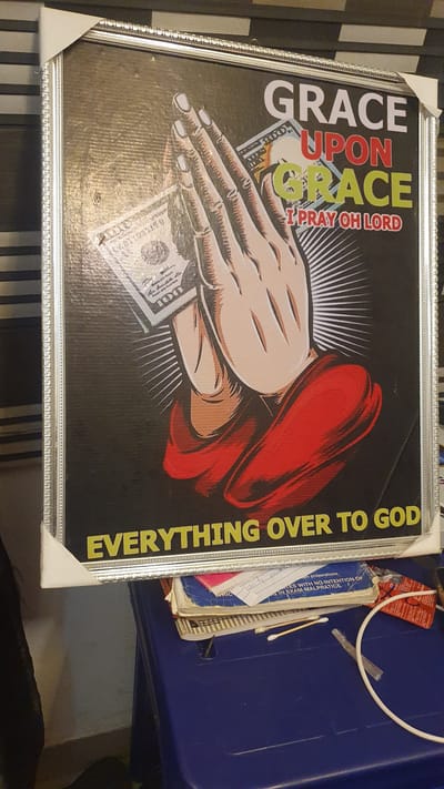 The image captures a framed piece of artwork placed on a striped surface, likely indoors in Awka, Nigeria. The artwork, predominantly black, features a stylized illustration of light-skinned hands in a praying or supplicating gesture. Several US hundred-dollar bills ($100) are visible beneath the hands, implying a connection between faith and prosperity. To the right of the hands, a vibrant red cap or headwear is depicted, with white rays emanating from behind the central figures.

Prominent text on the artwork includes "GRACE UPON GRACE" in large white and green letters, "I PRAY OH LORD" in smaller red text, and "EVERYTHING OVER TO GOD" in yellow letters running vertically along the right edge. The artwork is encased in a silver-colored frame with decorative patterns, and white protective corner pieces are still attached, suggesting it might be a newly acquired item.

Beside the framed artwork, a blue plastic container holds various miscellaneous items. Among these, several books or notebooks are discernible, along with a few Q-tips or cotton swabs, a white tube or cable, and a red tag. Partially legible text on one of the visible papers or books includes "am preparations," "TES WITH NO INTENTION OF," and "IN EXAM-MALPRATICE," suggesting academic or legal subject matter. The scene presents a personal setting, blending religious iconography with everyday items.