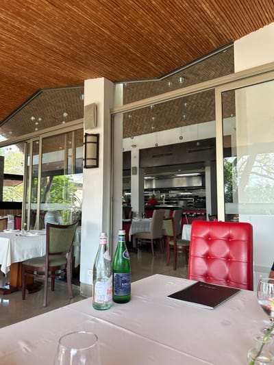 The image shows a partially covered outdoor patio area of a restaurant with a white tablecloth and a red leather chair. The restaurant has a wood slat ceiling, a white wall, and a large glass window that looks out onto a similar patio area. There are two glass bottles of water on the table. One is a "Pan" brand and the other is a "S.Pellegrino" brand. Two wine glasses sit on the table. One is empty and the other has a partially filled glass of wine. There is a closed black menu on the table. The location of the image is in Santa Ana, Costa Rica. The coordinates of the location are latitude 9.937, longitude -84.189. The environment appears to be a warm climate based on the outdoor patio area. The area appears to be well-lit. One person is visible outside the window, but their face is obscured by the glass. There are numerous empty chairs visible in the scene. The scene is a static one, with no visible action or interaction.