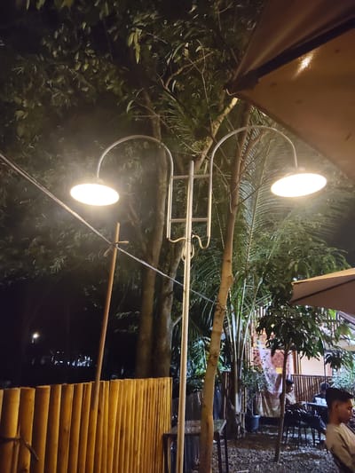 This image captures an outdoor seating area at night, possibly a cafe or restaurant, located in Glokaron, Indonesia. A double-headed street lamp illuminates the scene, casting a warm glow on the surrounding foliage and a portion of a tan-colored awning overhead. The ground is covered in gravel. In the foreground, a short, vertical fence made of bamboo poles is visible. In the background, several trees with lush green leaves are present, with some showing distinct palm fronds. Further back, a building with a decorative tapestry can be seen, and a few individuals are seated at tables, engaged in conversation or simply present in the space. One person in the right foreground is partially visible, looking towards the left.
