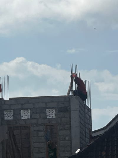 A person wearing a red shirt and dark pants is working on top of a building under construction in Sukawati, Indonesia. The building is made of grey concrete blocks, with rebar sticking out from the top. The sky is partly cloudy with a bird in the distance. The scene is captured from a low angle, looking up at the construction site.