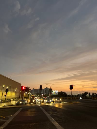 This image depicts a street scene in Arica, Chile, during twilight. The sky is a gradient of gray, pale orange, and yellow, with scattered clouds. In the foreground, a dark asphalt road shows lane markings and several vehicles, including cars and a truck, are visible. Traffic lights, some showing red signals, are positioned at intersections. The left side of the image features a building with illuminated windows and lampposts. In the background, a cluster of high-rise buildings is silhouetted against the sky, with some industrial structures and vegetation also present. A large billboard is visible in the middle ground. The scene suggests moderate traffic flow and the transition from day to night.
