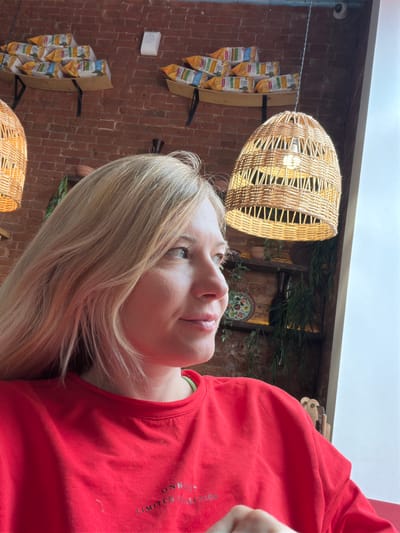 A medium shot, portrait-style image captures a woman with blonde hair in a red sweatshirt. She is facing right, looking away from the camera. The background appears to be an indoor setting, possibly a cafe or restaurant, with a brick wall, shelves displaying bags of snacks, and woven pendant lights. The image is taken from a slightly low angle.