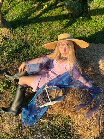 A young woman with blonde hair lies on her back, nestled between a bale of hay and a grassy area. She wears a large straw hat adorned with a floral crown of purple and green flowers. Her eyes are open, and she gazes upwards with a serene expression. She is dressed in a pale lavender dress and a translucent blue, shimmering overlay. On her right hip rests a woven basket filled with lavender sprigs. She is also wearing black, knee-high rubber boots. The scene is bathed in the warm, golden light of what appears to be late afternoon or early evening. The sun casts long shadows across the grass and hay. The location appears to be rural, possibly a field or meadow, given the presence of hay bales and natural vegetation. The overall mood is peaceful and ethereal.