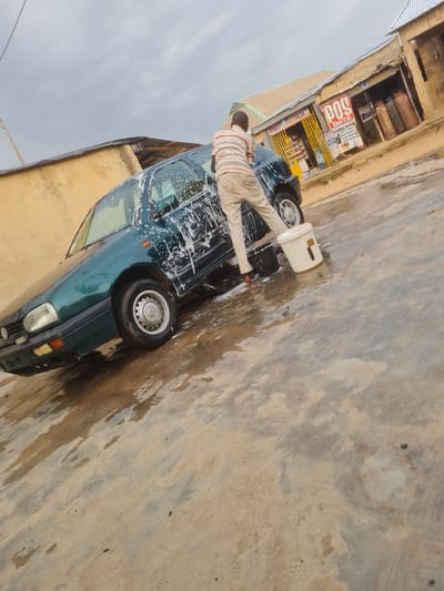 In Madalla, Nigeria, a man is washing a dark green Volkswagen Golf car. He is wearing a striped shirt and light-colored pants, and is using a white bucket to wash the vehicle, which is covered in soap suds. The car is parked on a wet, sandy surface, possibly a roadside car wash. In the background, there are several small shops with corrugated metal roofs, and some large gas cylinders are visible outside one of the shops. The sky is overcast.