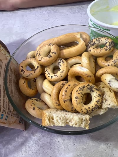 In the city of Bishkek, Kyrgyzstan, a clear glass bowl is filled with numerous circular bread products, some dusted with poppy seeds. To the right of the bowl, a white plastic container with a slightly opened lid holds a yellow substance, possibly butter or margarine. Partially visible on the left side of the frame is a brown paper package with text in Cyrillic. The background consists of a textured gray surface, suggesting a tabletop or counter. No individuals are clearly visible, but a blurred human limb is present in the upper portion of the image. The scene is likely indoors, given the lighting and arrangement of the objects.