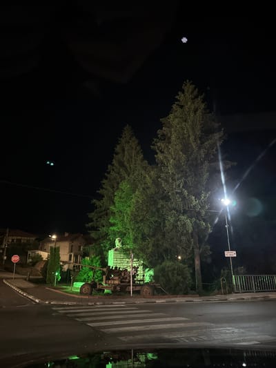 The image depicts a street scene at night in Barziya, Bulgaria. A large, leafy tree dominates the center of the frame, illuminated by a street lamp overhead. To the right of the tree is a street with a pedestrian crossing. There is a dark object that looks like a derelict farm machine or trailer standing beside the street, partly obscured by the tree and lighting. A street sign is visible next to the street lamp. In the lower right of the image, buildings are visible, along with a stop sign. The sky is dark, with one or two lens flares visible. The overall scene has a somewhat eerie feel due to the dark lighting and the presence of the derelict machinery.