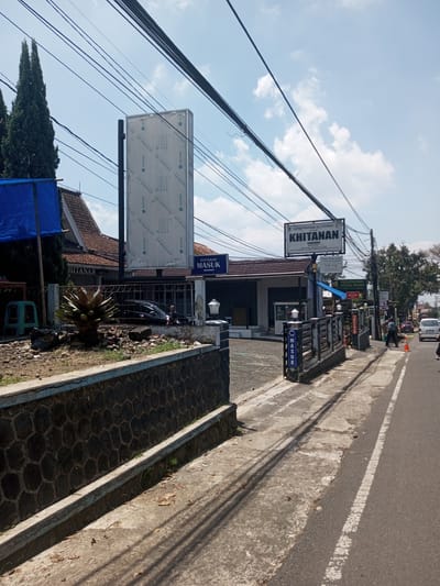 The image depicts a street scene with a medical clinic specializing in circumcision ("KHITANAN"). The clinic, "KLINIK PRATAMA AL-FITHRAH," has a prominent sign and an entrance marked "MASUK" (Enter). There is a tall, blank sign next to the clinic. Power lines are visible overhead. The scene includes a road with a white line marking the edge, a sidewalk, and a low stone wall bordering the property. A person is walking down the sidewalk, and cars are parked or driving on the road. The location of the scene is in Indonesia, due to the language on the signs.