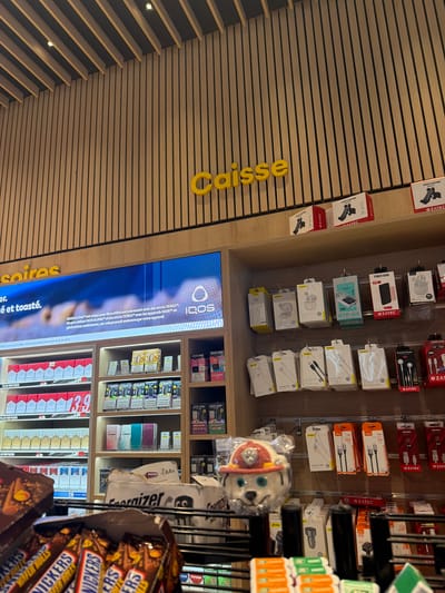 A retail space in El Mansouria, Morocco features wooden slat walls with the yellow word "Caisse" and displays various products. The displays include phone accessories by OZATEC, electronic cigarettes with IQOS branding, and shelves stocked with cigarettes, e-cigarettes from Geek Bar and candy bars such as Snickers.