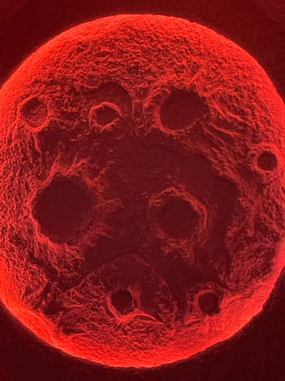 The image displays a highly magnified, irregularly spherical object, predominantly rendered in shades of red and dark red against an unidentifiable black background. The object's surface is heavily textured, appearing rough, porous, and undulating, with numerous depressions, pits, and crater-like formations of varying sizes. Brighter red highlights illuminate the raised contours and edges, particularly on the upper and left sides of the sphere, suggesting a directional light source. Deeper recesses and shadowed areas are depicted in darker crimson tones, approaching black. The object occupies the majority of the frame, presented in extreme close-up. No people, other distinct subjects, objects beyond the primary sphere, actions, or interactions are visible. The surrounding environment is an undifferentiated void. The image provides no visual information to determine its geographic location, including within the city of Vinnytsia, Ukraine.