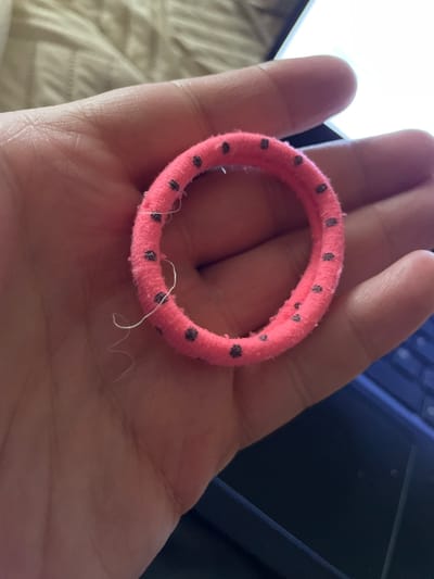 A person's hand, with visible skin texture and lines, holds a pink, fuzzy scrunchie with dark polka dots. A single white thread is dangling from the scrunchie. The hand is positioned over a dark surface, possibly a laptop keyboard, with a sliver of a white object in the upper right corner, suggesting an indoor setting. There is no information in the image that could identify its location in Ivano-Frankivsk, Ukraine.
