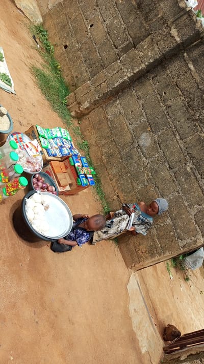 In Kagarko South, Nigeria, a young child is seated on the ground next to a large silver tray containing white food items. The child is wearing a blue and white patterned garment. Beside the tray, a woman, dressed in a blue and white patterned outfit and a grey head covering, is seated and appears to be writing in a notebook. The ground is a reddish-brown dirt. In the background, there are various items for sale, including packaged snacks, drinks in plastic bottles, and red onions in a dark bowl. A rough, textured stone wall forms the backdrop to the scene.