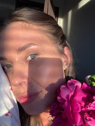 This is a close-up, sideways shot of a young person's face and upper body, partially obscured by a large, vibrant pink peony flower. The person has fair skin, light brown hair, and striking green eyes. They are wearing several small earrings, including a silver hoop in the upper ear and a diamond stud and a square-shaped stud in the lower ear. A delicate silver necklace with a circular pendant is visible around their neck.
The scene is bathed in natural light, casting strong shadows across the person's face, suggesting it is either early morning or late afternoon. The light creates a dramatic effect, highlighting the texture of their skin and the delicate features of their face. The background is dark and out of focus, with some lighter areas that might indicate a wall or indoor setting. The person is lying down, their head resting on a white fabric that appears to have a subtle cherry pattern. The overall mood is peaceful and serene, with a touch of natural beauty emphasized by the flower. The location is identified as Chernushka, Russia.