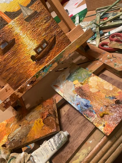 The image shows an artist's workspace. An unfinished oil painting depicting a building is on an easel. Several paint palettes covered in oil paints, including yellows, browns, blues, and greens, are visible. A tube of Winsor & Newton Winton oil color (Titanium White) rests near a palette. Brushes and art supplies are in the upper left corner. The scene is likely in an indoor studio. There is no indication of location in the city of Maidstone, United Kingdom.