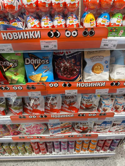 The image shows a shelf in a store, likely a supermarket, displaying a variety of candy and snack products. The products are arranged in rows and include items such as Kinder Joy, Doritos, and various chocolate bars and cookies. The shelves are well-lit, and the products are neatly presented. There are price tags and labels visible in Russian and possibly other languages. A prominent orange display strip with cartoon eyes is placed across the shelves, indicating new products or promotions. The overall impression is a typical retail environment for confectionery and snacks. The location is noted as Родниковское сельское поселение, Ukraine.